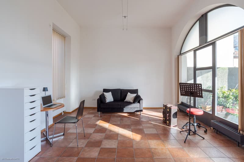 Spacious living room with large arched window, hardwood tile flooring, black sofa, desk with laptop, and music equipment