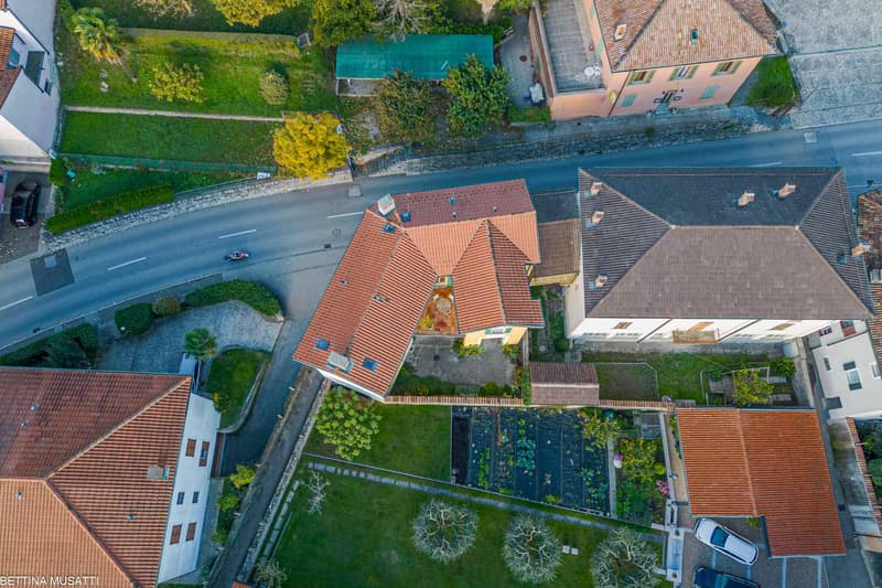 The image shows an aerial view of a residential area with a mix of buildings, including houses and apartment buildings. The roofs are predominantly made of red tiles, and the buildings are surrounded by lush greenery, including trees and well-maintained g