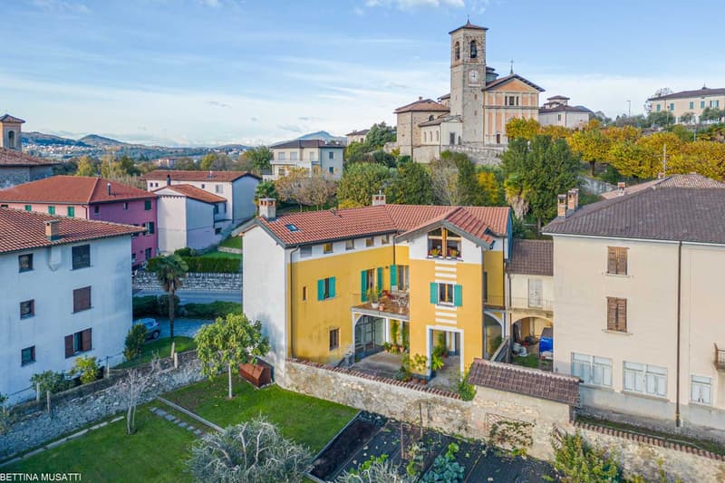 The image shows a picturesque town with colorful buildings, a church tower, and lush greenery. The buildings have red tile roofs and some have balconies or terraces. The town is situated on a hillside, offering views of the surrounding landscape.