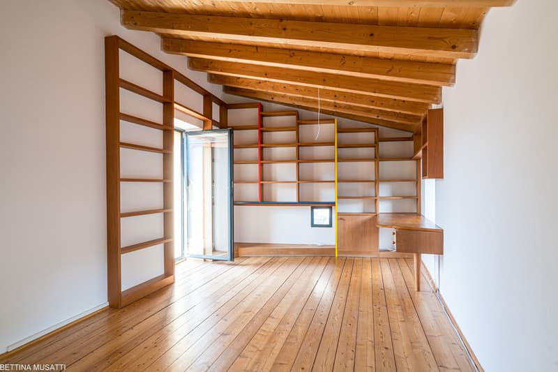 Spacious room with wooden beamed ceiling, wooden shelving units along the walls, and a wooden floor. The room has a large window that provides natural light and access to a balcony or terrace.