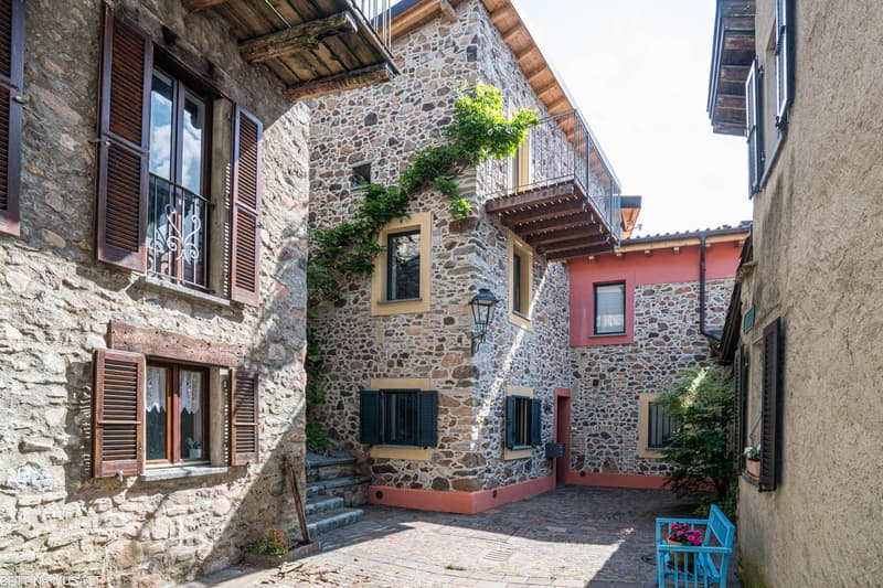 This image shows a traditional stone building with wooden shutters and balconies. The building has a rustic, Mediterranean-style architecture with a cobblestone pathway leading to the entrance. There are potted plants and a small bench visible, adding to