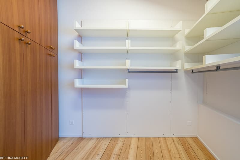 The image shows a bedroom with a wooden closet and built-in shelves along the wall. The room has a hardwood floor and a clean, minimalist design.