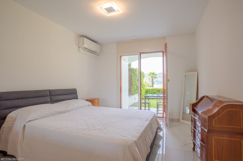 Spacious bedroom with a large bed, gray sofa, wooden dresser, and sliding glass door leading to a balcony or terrace with a view of the surrounding greenery.