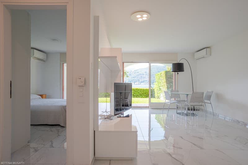 Open-plan living area with dining table and chairs, TV stand, and sliding glass doors leading to a balcony or terrace with a view of the surrounding greenery. The bedroom is visible through an open doorway. The apartment features modern, minimalist decor