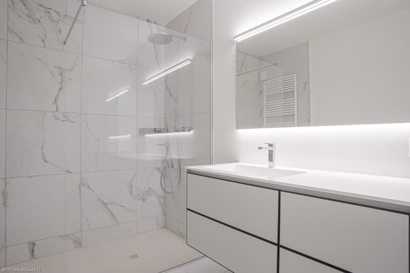 Spacious bathroom with marble-like tiles covering the walls and floor, a large walk-in shower with a rainfall showerhead, a vanity unit with a sink and drawers, and a backlit mirror above the vanity. The room is well-lit with recessed lighting.