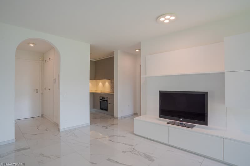 Spacious open-plan living area with white walls, marble-style tile flooring, recessed lighting, and a built-in TV cabinet. The kitchen area is visible through an archway, featuring white cabinetry and a built-in oven or dishwasher.