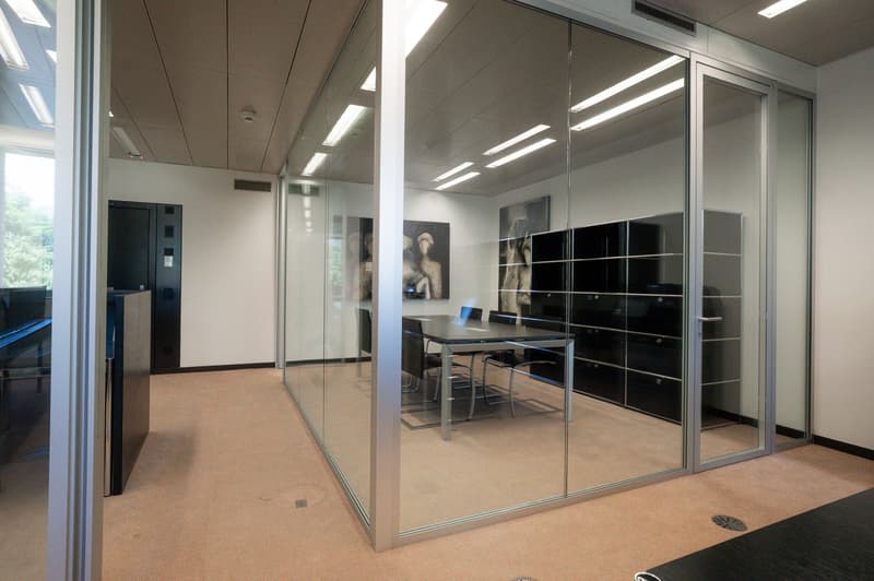 modern office room with glass walls and partitions, beige floor, table and chairs, black storage units, 3 paintings on the wall