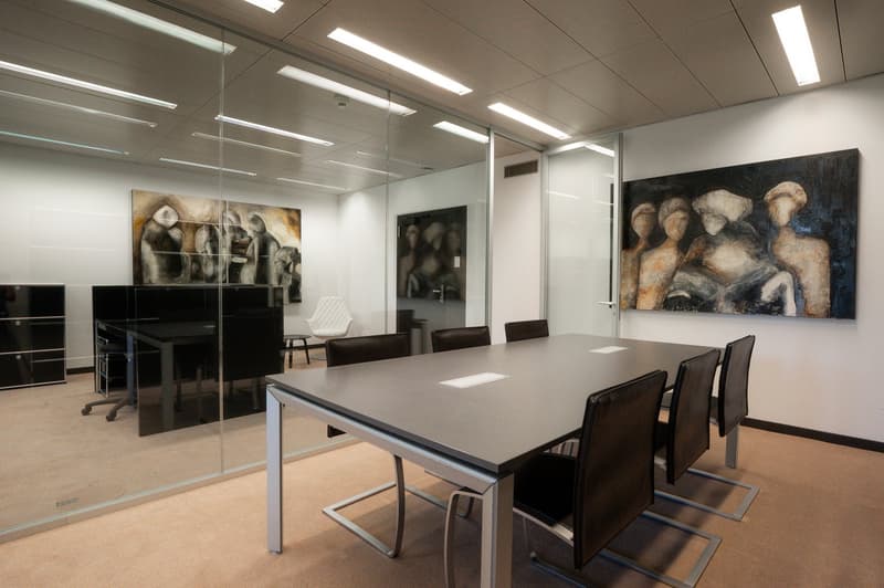 Modern office, large glass walls, wooden flooring, long table, black leather chairs, paintings on the wall