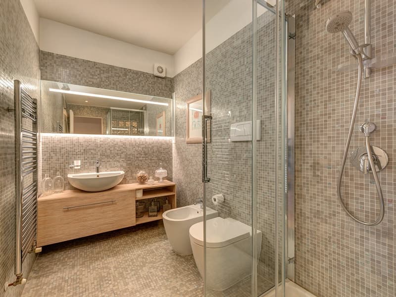 Compact bathroom with modern fixtures, tiled flooring and walls, glass walk-in shower, wooden vanity, white toilet, bidet, illuminated mirror, and towel warmer.