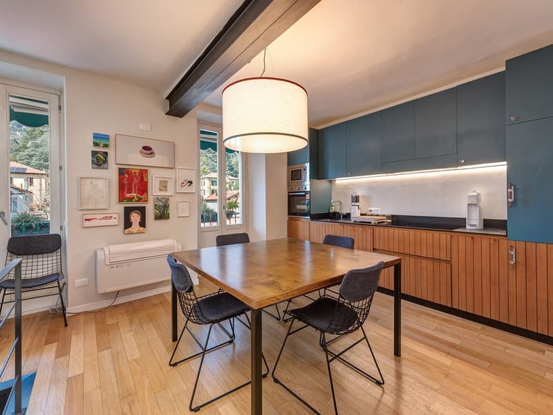 Dining area, wooden floor, wooden table, 4 chairs, kitchen area with black countertop, white appliances, cabinets, artwork on wall, wooden cabinets, pendant light.