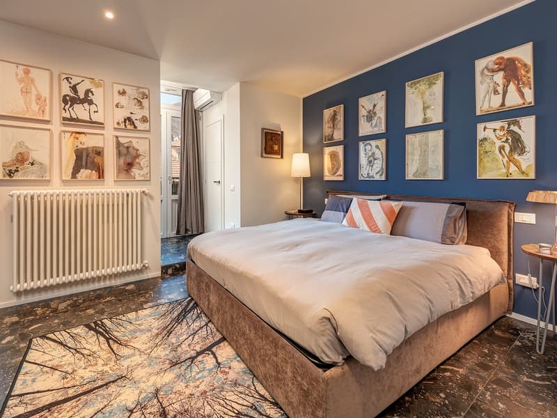 Master bedroom: double bed, carpet, artwork on the walls, table with lamp, white radiator, tiled floor, air conditioning, open doorway