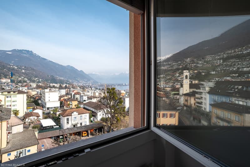 Cityscape with buildings and mountains, reflection of building in the window, a town center with buildings, houses, a church with a bell tower, mountains, hills, a river, trees, and a blue sky