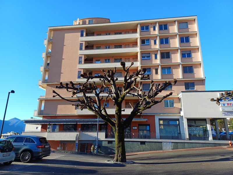 Multi-story residential building with beige walls and glass windows, multiple balconies on each floor, small trees, metal fences in front of the ground floor, cars parked on the street.