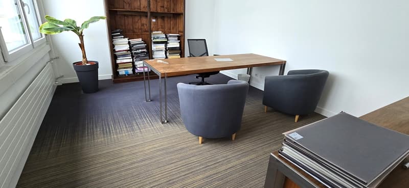 Desk with office chair, 2 additional chairs, wooden bookcase, stack of books, large window, potted plant, carpeted floor, radiator