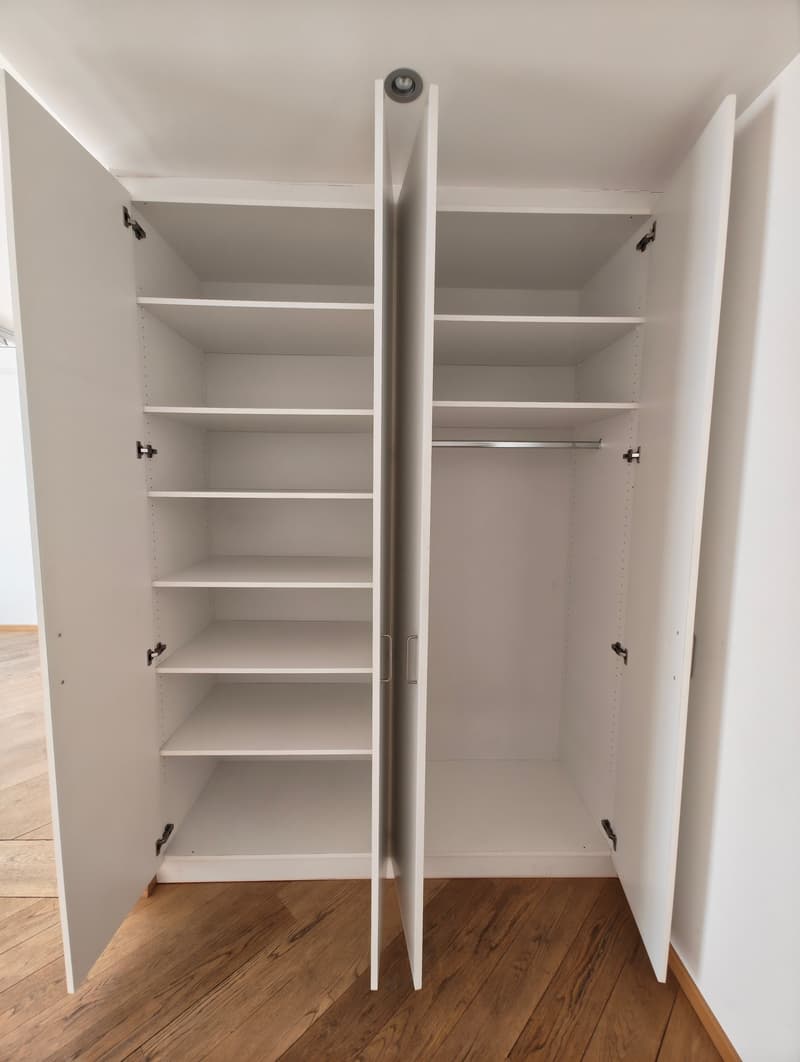 Large white wardrobe with multiple shelves and a hanging rail, fully open. Wooden floor.