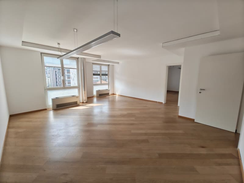 Empty room, wooden floor, white walls, large windows, blinds, recessed ceiling lights, two radiators under the windows, no visible features.