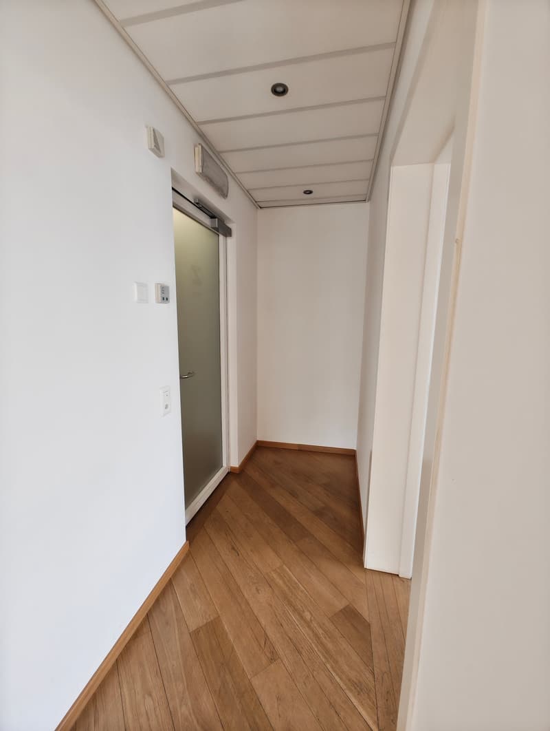 Empty hallway with a closed door, wooden floor, white walls, and ceiling-mounted lights