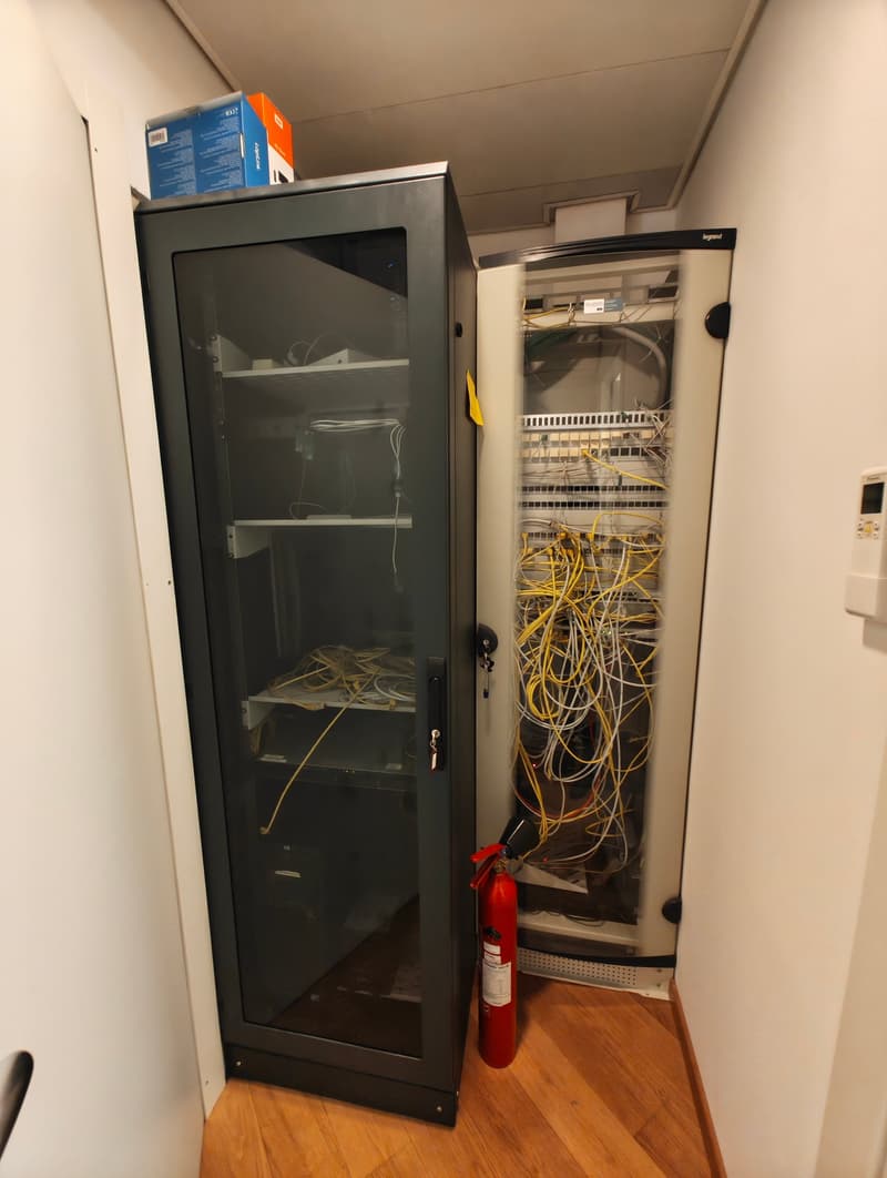 Metal cabinet with glass door, electrical equipment inside, yellow cables, red fire extinguisher, white walls, wooden floor