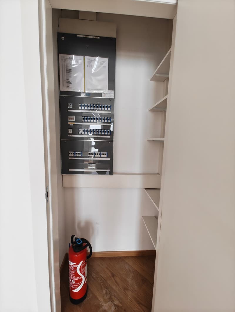 Small room with electrical panel on wall, three shelves, fire extinguisher on wooden floor