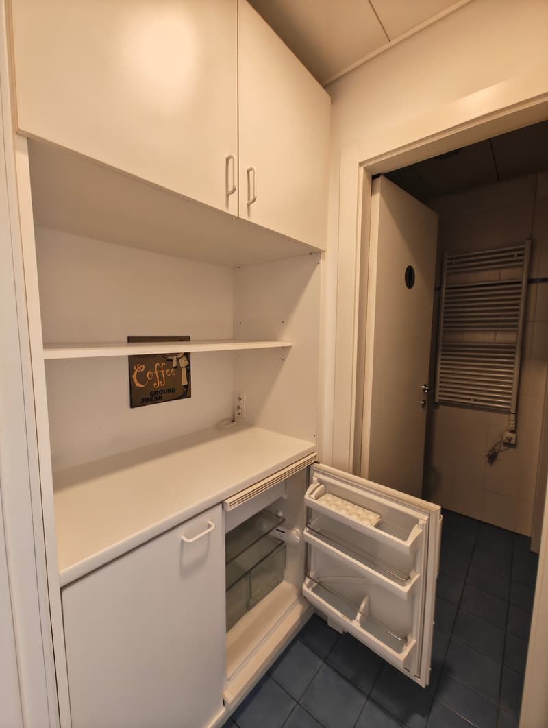 Clean, white kitchen with tiled flooring, shelves, a counter with an open fridge, power outlet, and a coffee ground sign on the middle shelf. An open door leads to an adjacent room with a radiator. The room has white walls and a white ceiling.