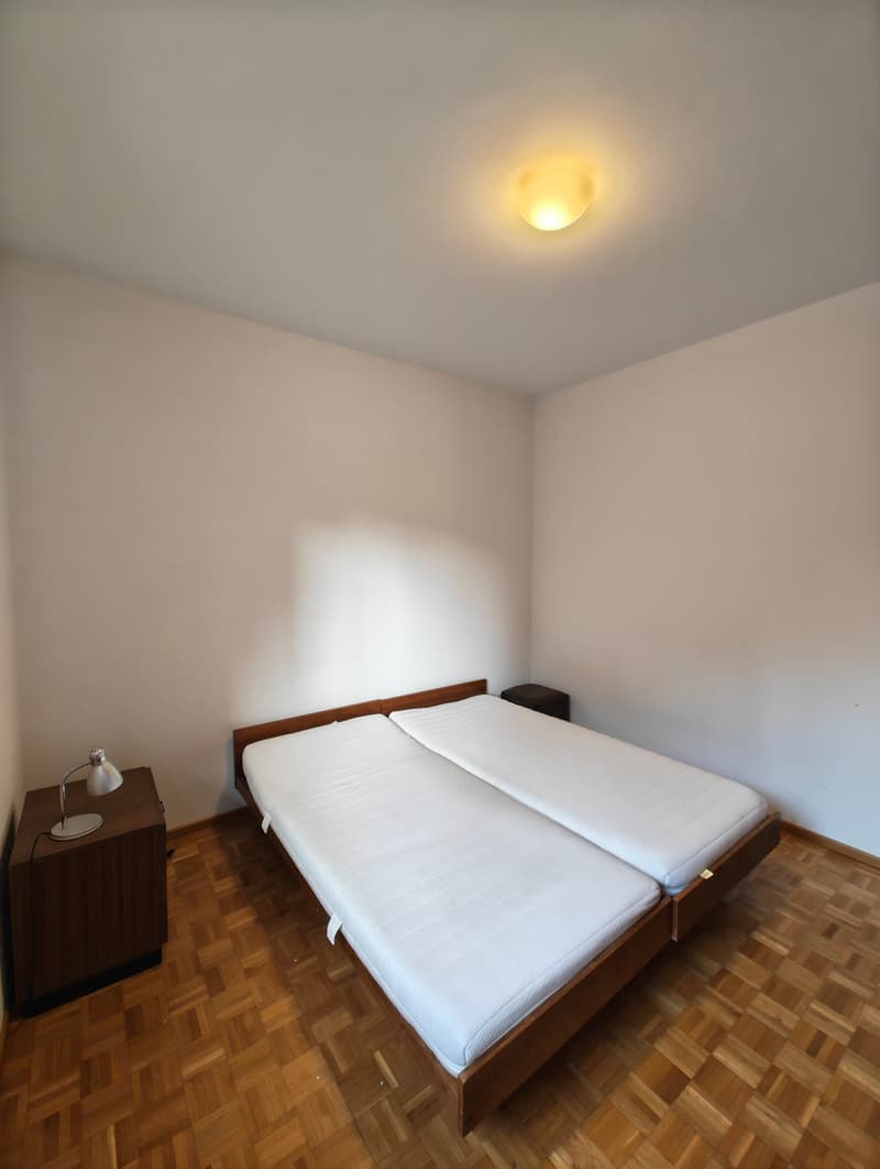 Bedroom with two single beds, wooden floor, white walls, ceiling lamp, nightstands with lamps
