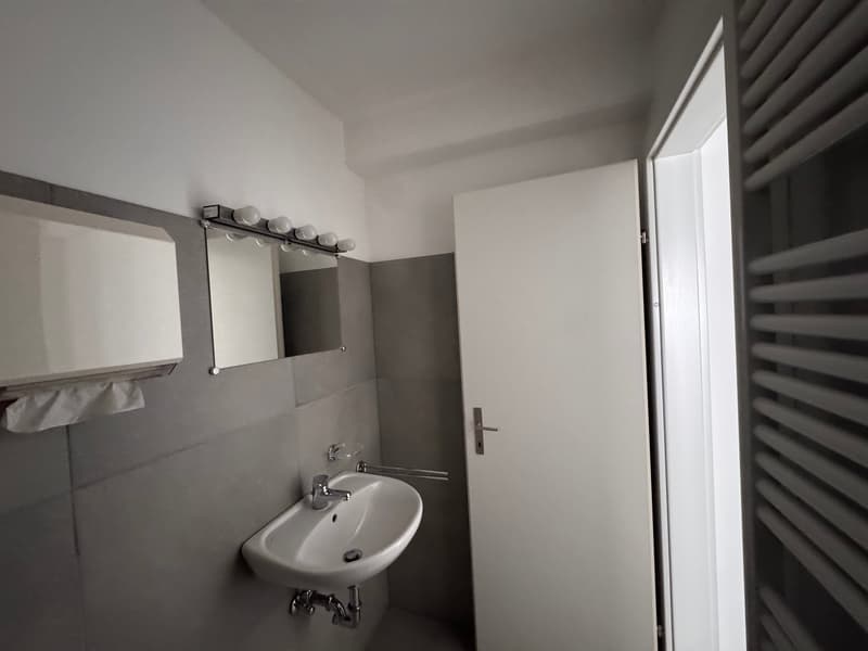 white washbasin, mirror with mounted lights, white door, grey tiles, towel rack