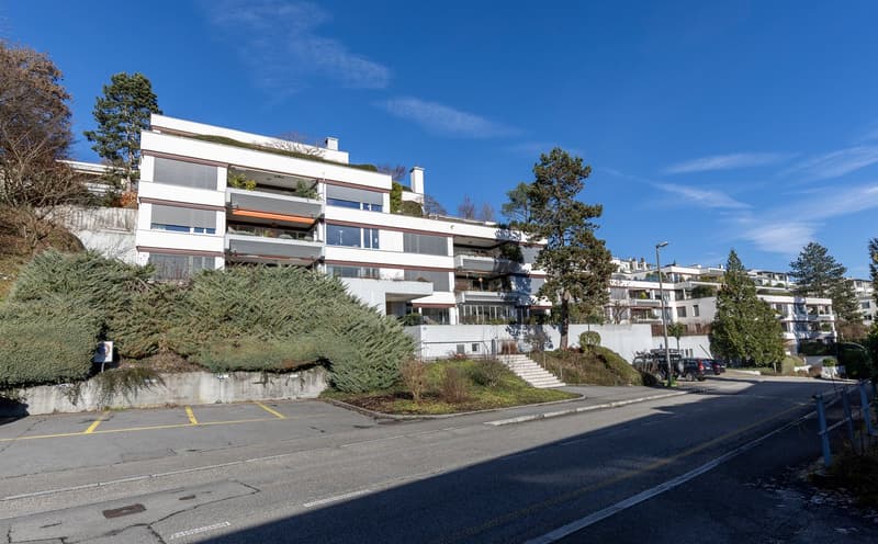 Modern multi-story building with balconies and parking area on a sloped terrain.