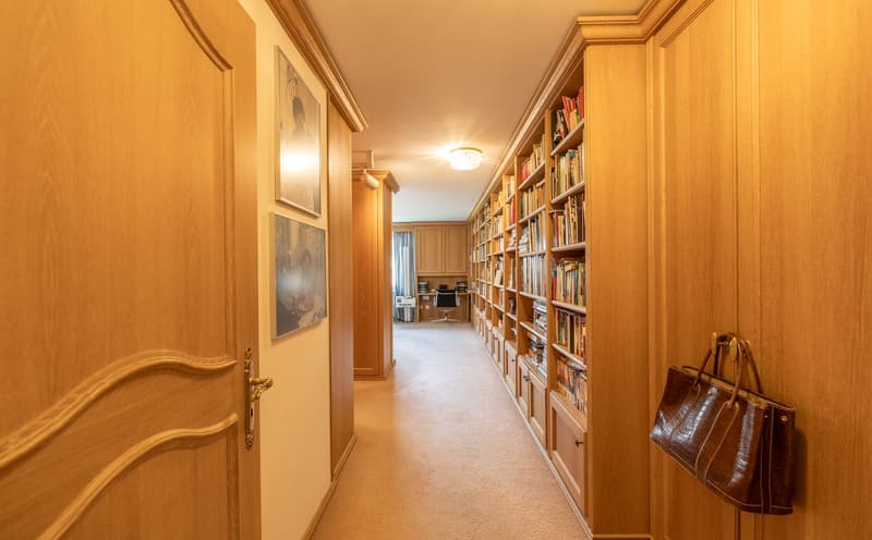 A hallway with wooden doors, carpeted floor, and built-in bookshelves filled with books. A handbag is hanging on the wall.