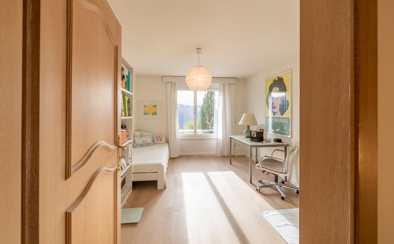 A small bedroom with a bed, desk, and chair. The room has a bookshelf, a poster, and a lampshade. The bed has a pillow and a rug. The desk has a lamp, a computer, and a book. The room has a window with curtains and a view of a tree.