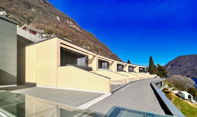 A modern multi-story building with a brick facade, large windows, and a balcony overlooking a mountainous landscape with a clear blue sky.