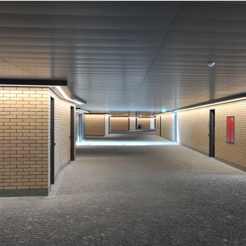 Long, well-lit hallway with brick walls, a tiled floor, and multiple doors leading to other rooms or areas. The ceiling has a series of parallel beams running across it, providing a modern and industrial aesthetic.