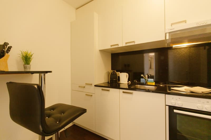 Compact kitchen with white cabinets, black countertops, and a dishwasher. There is a small table with a chair in the corner.