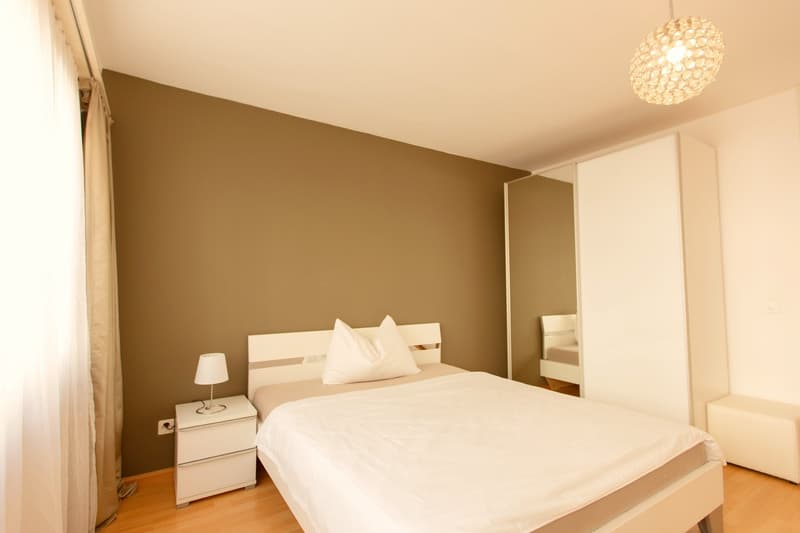 Spacious bedroom with beige walls, white bed with pillows, nightstand with lamp, and a crystal chandelier overhead. There appears to be a balcony or terrace visible through the window.
