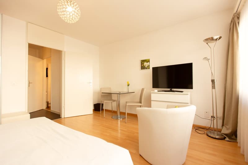 Spacious bedroom with hardwood floors, white walls, a large TV mounted on the wall, a small dining table with two chairs, and a white sofa. The room is well-lit with a crystal chandelier overhead.