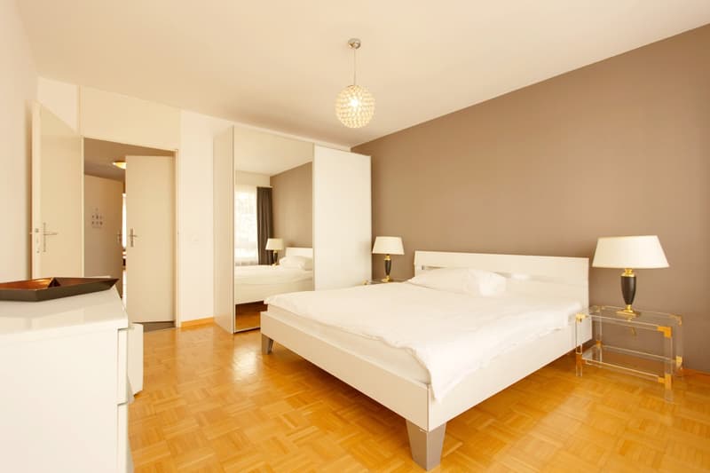 Spacious bedroom with white walls, hardwood floors, a large bed, bedside tables with lamps, and a crystal chandelier. There is a closet and a doorway leading to another room.