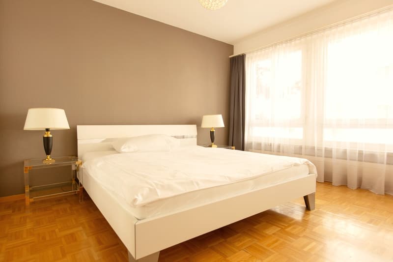 Spacious bedroom with white walls, large windows, hardwood floor, white bed frame, two bedside tables with lamps, and curtains covering the window