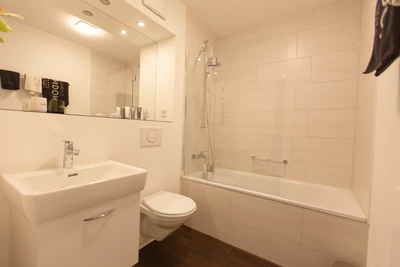 Modern bathroom with white tiles, a vanity sink, a toilet, and a bathtub with a shower curtain. There is a shelf above the vanity with some toiletries and a towel rack.