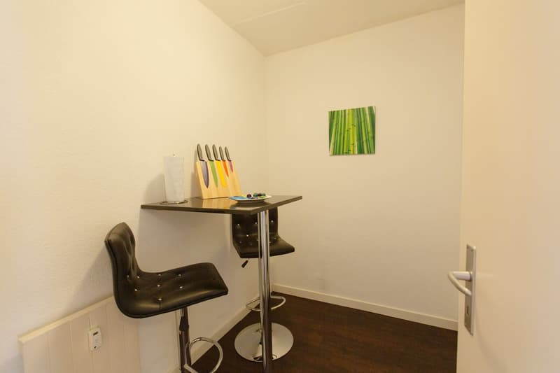 Small kitchen area with a wooden counter, a knife block, and a black adjustable chair. There is also a framed artwork of bamboo on the wall.