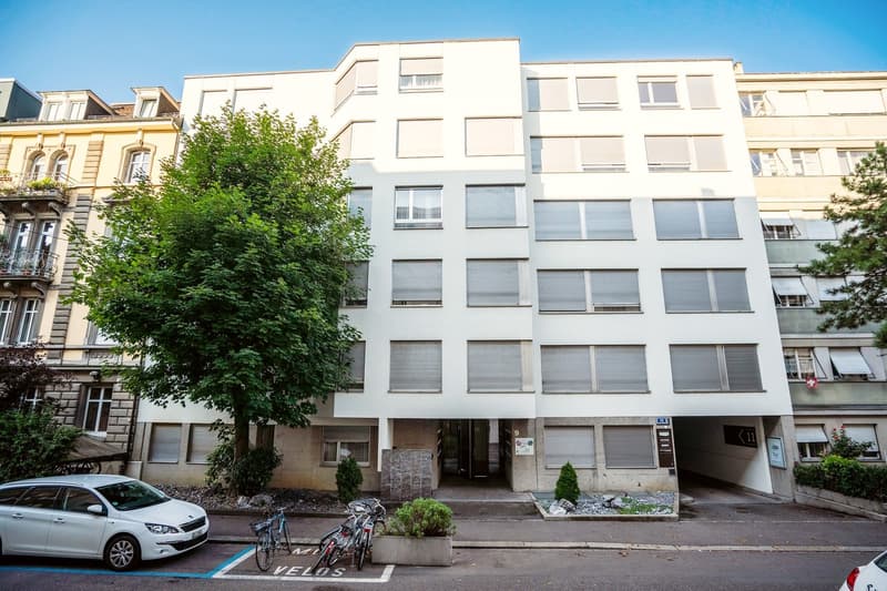 A multi-story apartment building with a modern, white facade. The building has several floors and windows, and there are trees and other buildings visible in the surrounding area. There is a car parked in front of the building and some bicycles parked nea