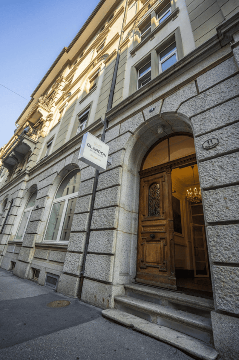 A multi-story building with a grand, ornate entrance. The building has a stone facade with large windows and a wooden door. The entrance is accessed by a set of stairs leading up to the doorway, which is adorned with decorative architectural elements. The