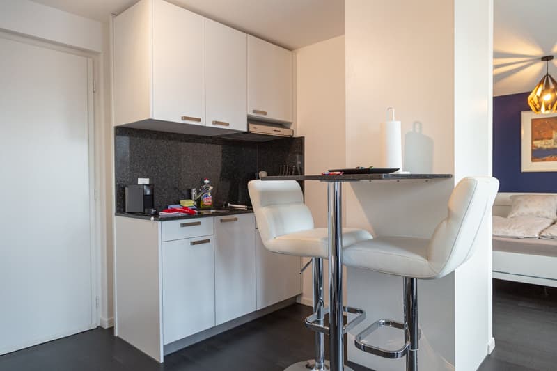 Compact kitchen with white cabinets, black granite countertop, built-in dishwasher, and bar seating area with two white leather stools