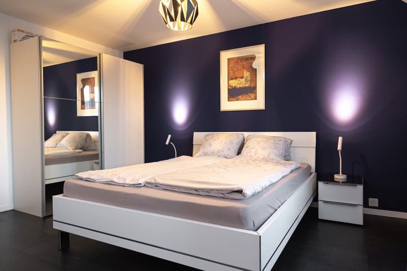 Spacious bedroom with a modern, minimalist design. The room features a large bed with white bedding, a built-in wardrobe, and a decorative lighting fixture. The walls are painted in a deep purple color, creating a cozy and sophisticated atmosphere. A fram