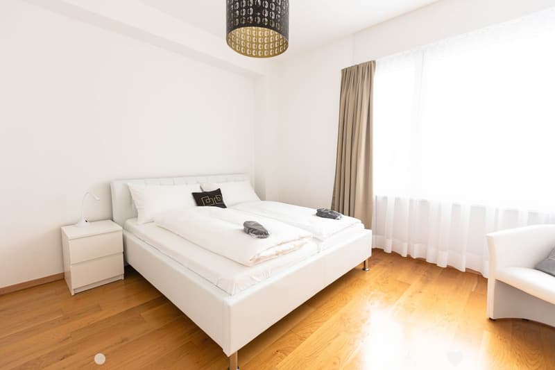 Spacious bedroom with white walls, hardwood flooring, a large bed with white bedding, a nightstand, and a patterned pendant light fixture. The room also features a curtained window, suggesting the presence of a balcony or terrace.