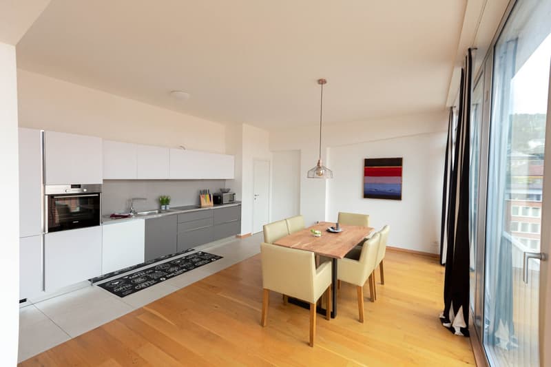The image shows a modern, open-plan kitchen and dining area. The kitchen features white and gray cabinets, a built-in oven, and a dishwasher. The dining area has a wooden table with beige chairs. The room has large windows, hardwood floors, and a pendant