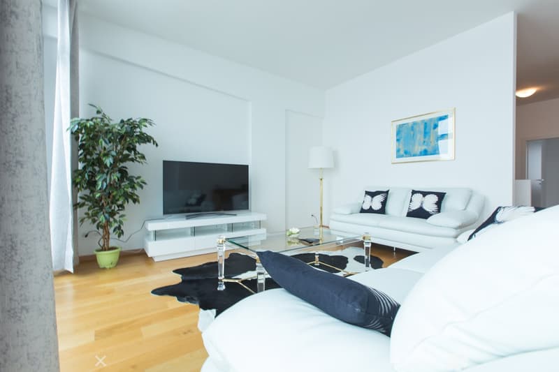 Spacious living room with white walls, hardwood floors, large windows, white sofa, glass coffee table, TV stand, and a potted plant.