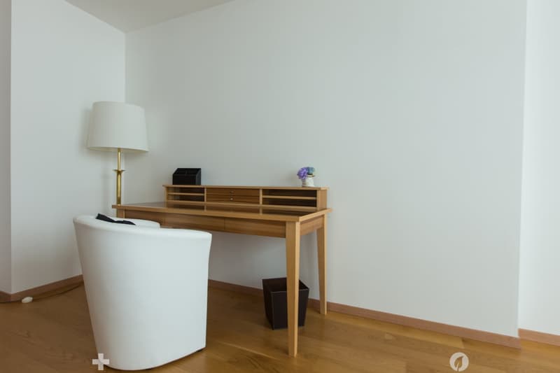 The image shows a minimalist and modern living room or study area. The room has white walls and a wooden floor. There is a wooden console table or desk with drawers, and a white armchair. A table lamp is placed on the console table, and a small vase with