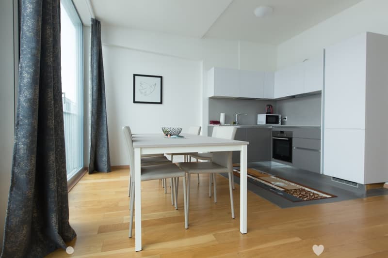 The image shows a modern, open-plan kitchen and dining area. The kitchen features grey cabinets and appliances, including a dishwasher. The dining table is a simple white rectangular table with four chairs. The room has large windows providing plenty of n