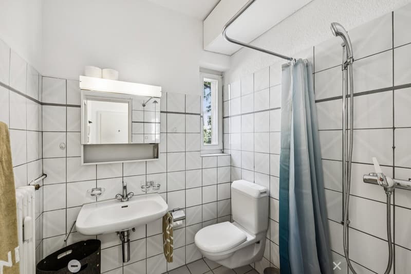 Tiled bathroom with a sink, toilet, and shower with a curtain. There is a mirror cabinet above the sink and a towel rack.