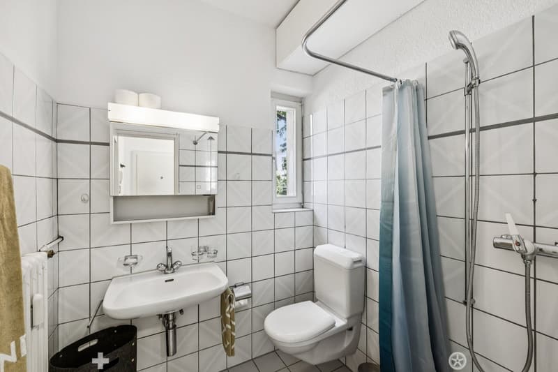 Tiled bathroom with a sink, toilet, and shower with a curtain. There is a mirror cabinet above the sink and a towel rack.