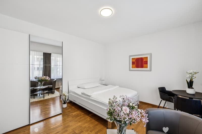 Spacious bedroom with hardwood floors, white walls, large window, bed, nightstand, artwork, and a vase of flowers on a table.